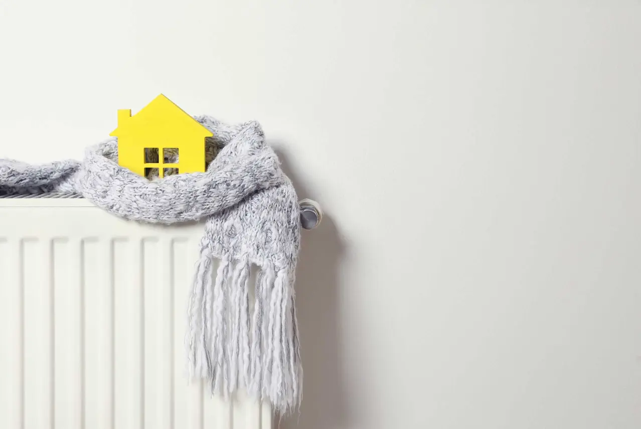 Machetă galbenă de casă înfășurată într-un fular gri, așezată pe un calorifer – simbol al termoizolării